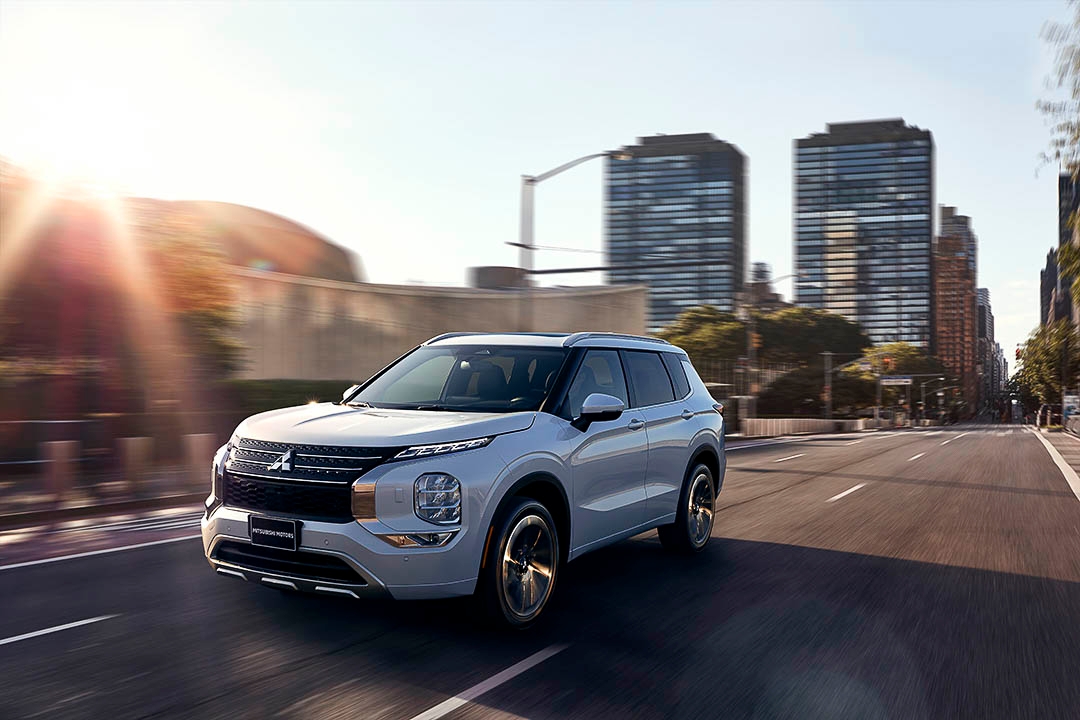 Mitsubishi Outlander 2022 blanc circulant en ville au coucher du soleil, avec immeubles modernes en arrière-plan.