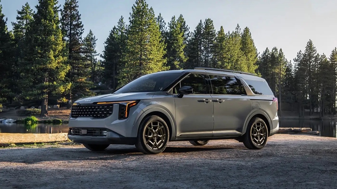 Kia Carnival 2025 gris stationnée en pleine nature près d’un lac, avec une forêt dense en arrière-plan, vue trois quarts avant.