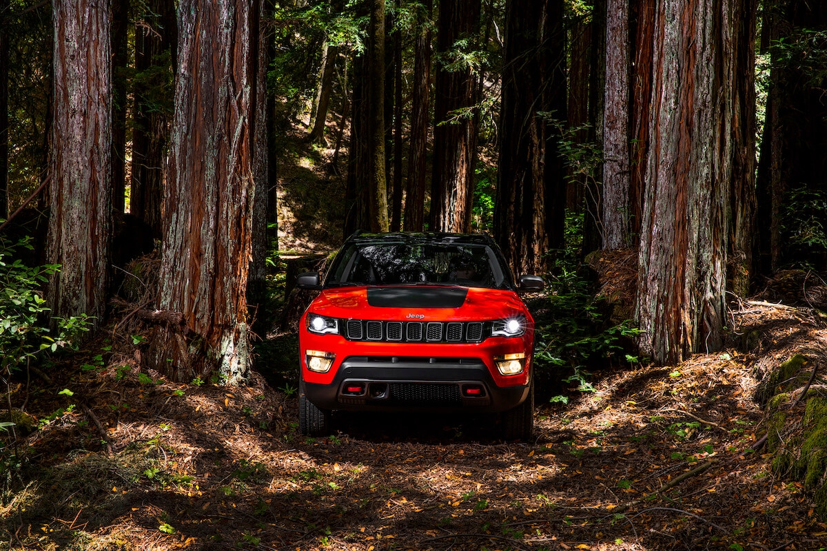 Jeep Compass 2021 rouge stationné en pleine forêt de conifères, vu de face avec phares allumés.
