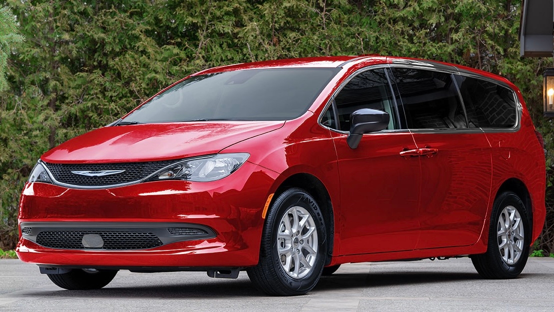Chrysler Grand Caravan 2025 rouge stationnée devant une haie de cèdres, vue trois quarts avant.