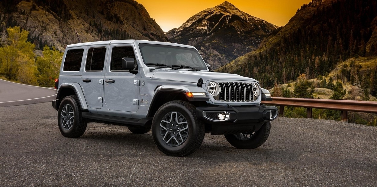 Jeep Wrangler 2025 argenté stationné sur une route de montagne sinueuse au coucher du soleil.