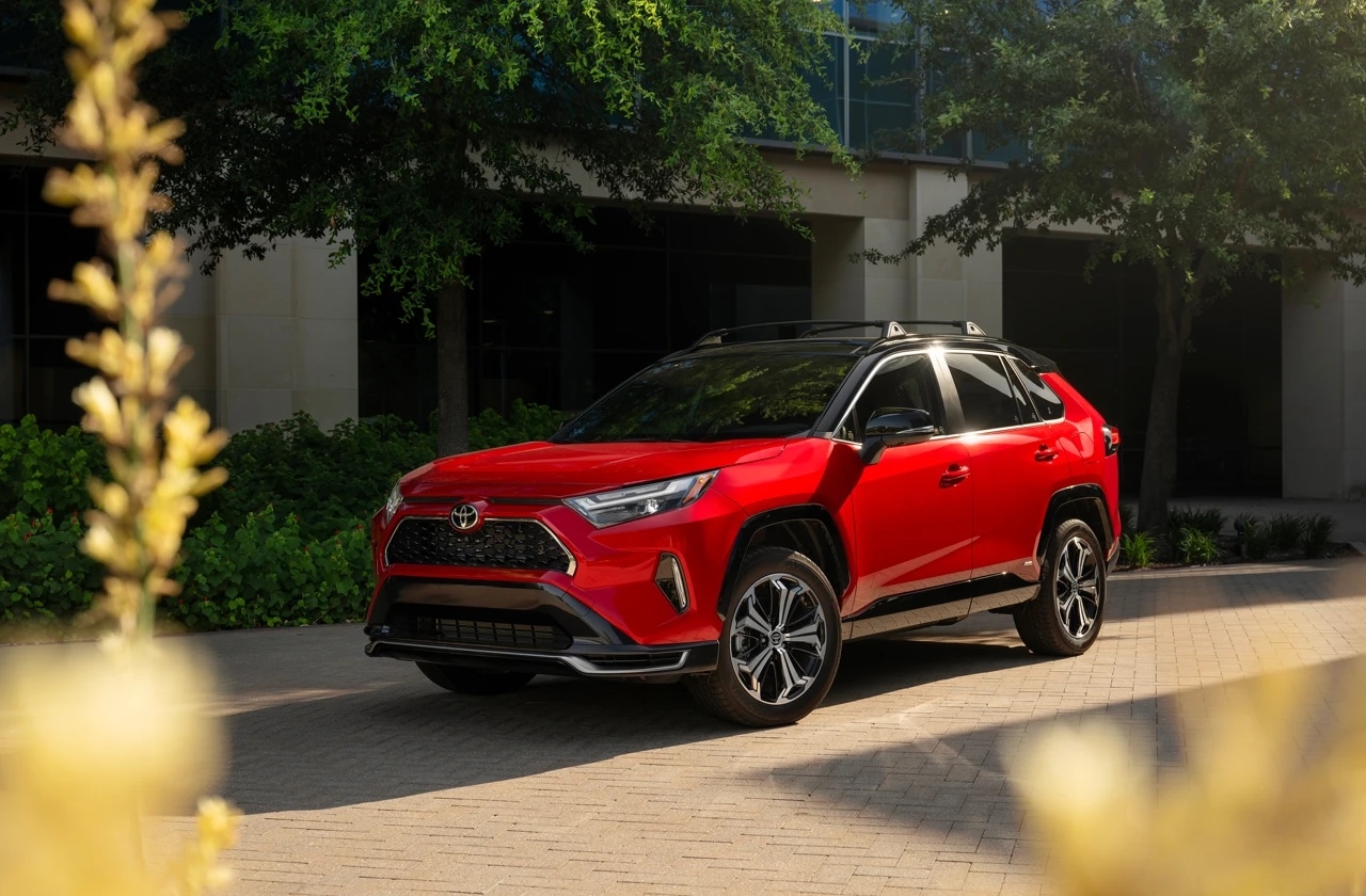 Toyota RAV4 hybride rechargeable 2025 rouge avec toit noir, garé dans un environnement urbain avec verdure.