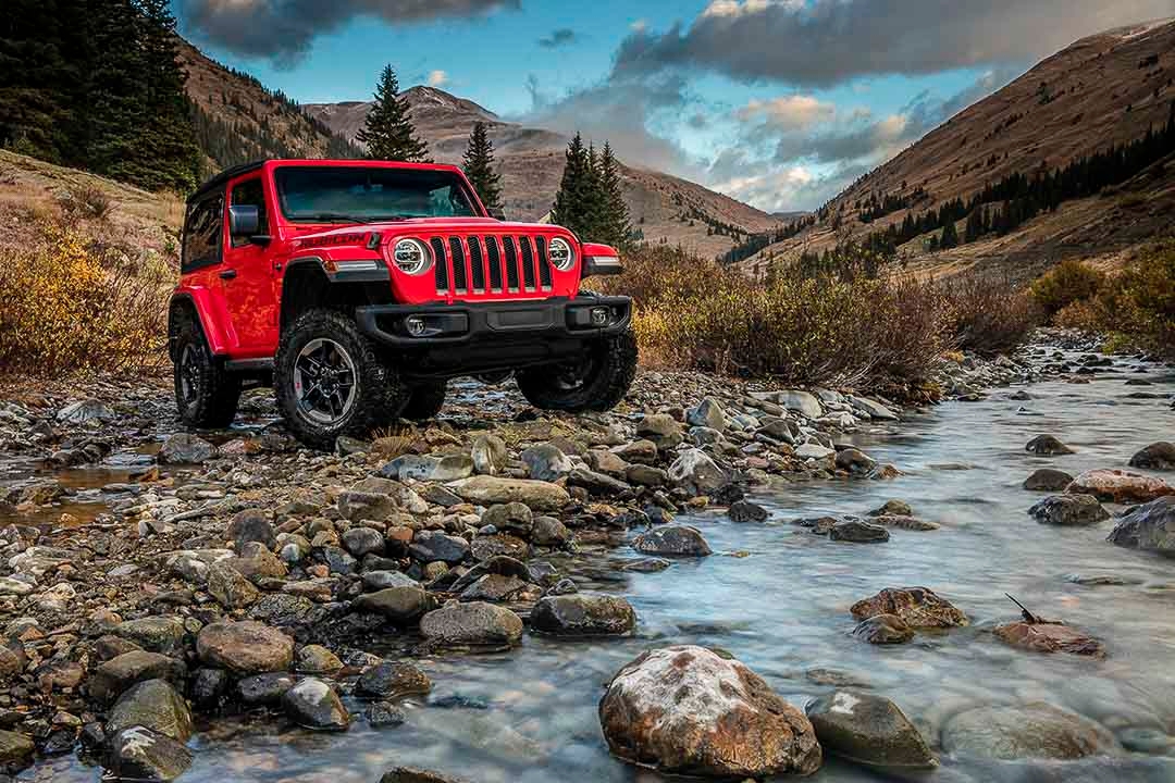 Jeep Wrangler Rubicon 2021 rouge sur un terrain rocheux près d’un ruisseau, en pleine nature montagneuse.