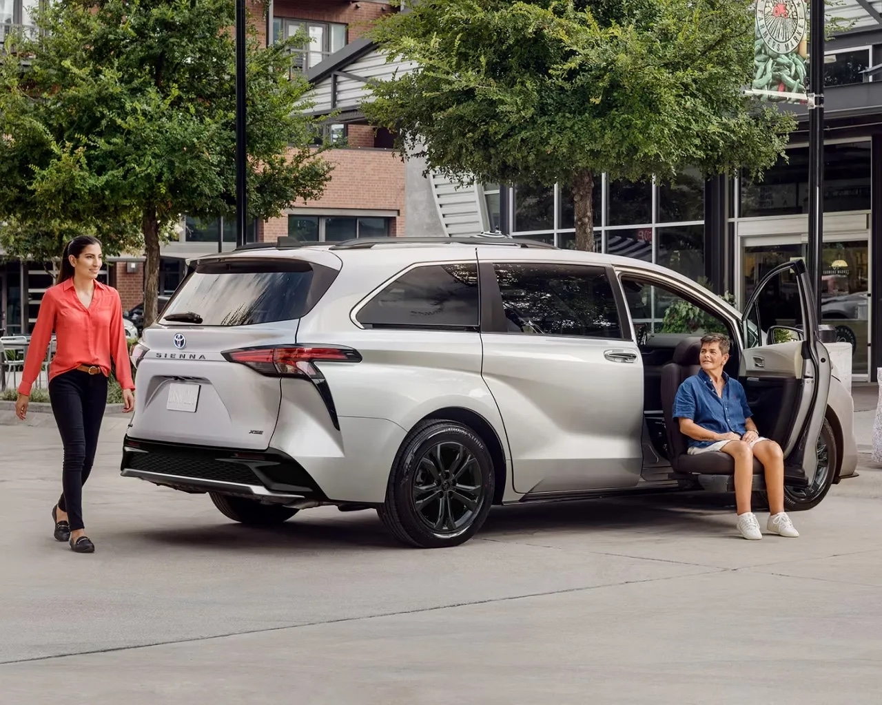 Toyota Sienna hybride 2025 argent avec siège pivotant déployé et une femme à l’extérieur, scène urbaine en arrière-plan.