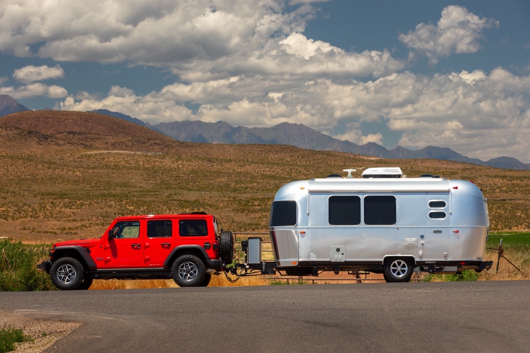Vue latérale d’un Jeep Wrangler Rubicon rouge 2025 attelé à une roulotte Airstream, paysage montagneux en arrière-plan.
